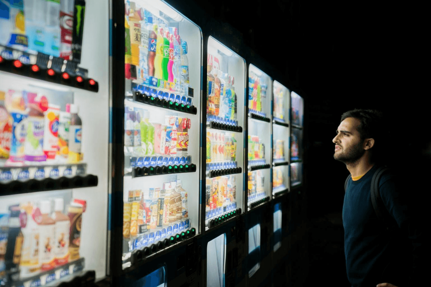 A person stands in front of brightly lit vending machines, gazing at an array of snacks and drinks. The scene is set at night, highlighting the contrast between the lit machines and the dark surroundings.