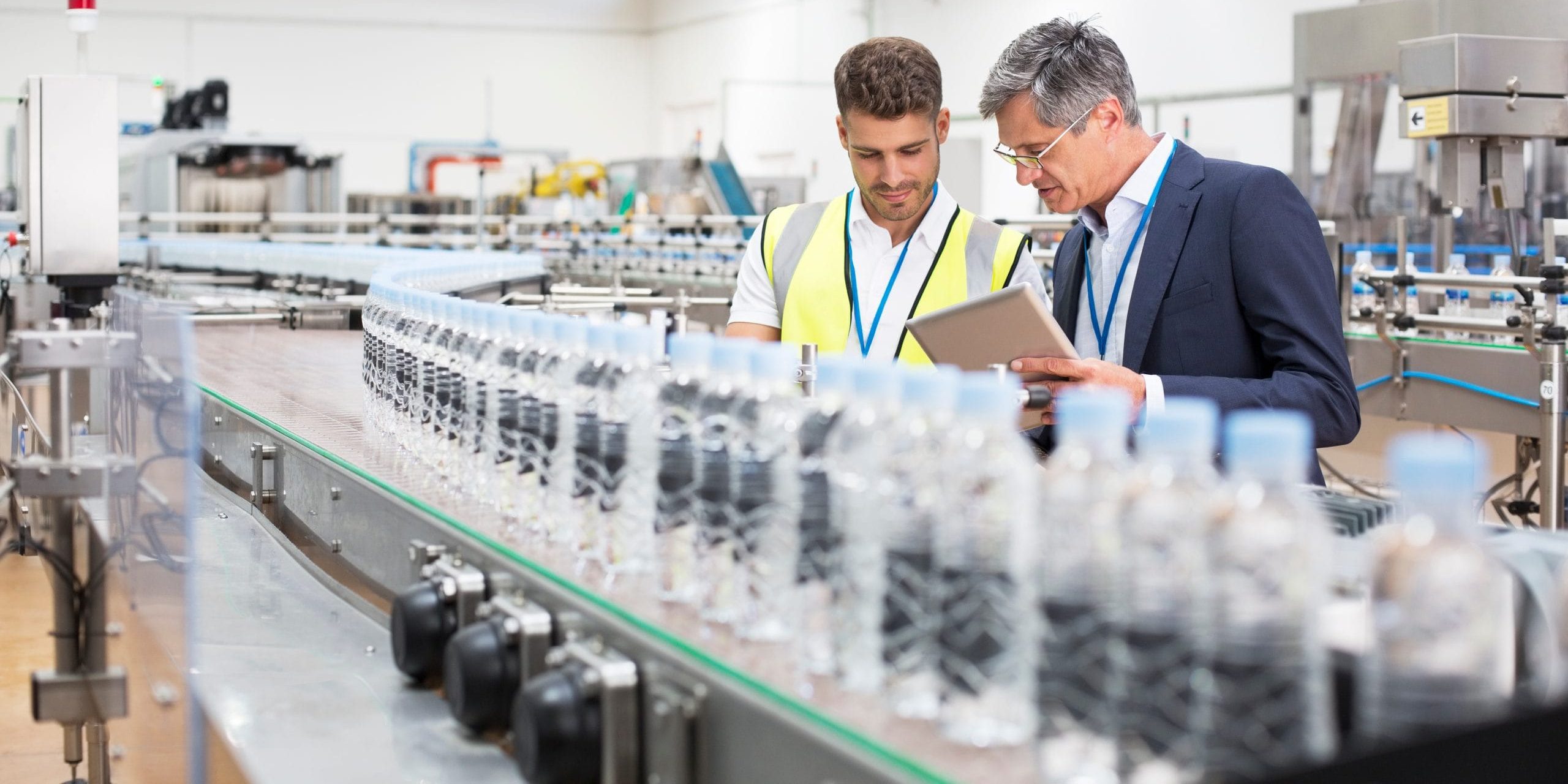 Two men in safety vests and lanyards discuss optimizing beverage production lines on a tablet in a modern bottling factory. They stand beside a conveyor belt with plastic water bottles, in a well-lit industrial setting.