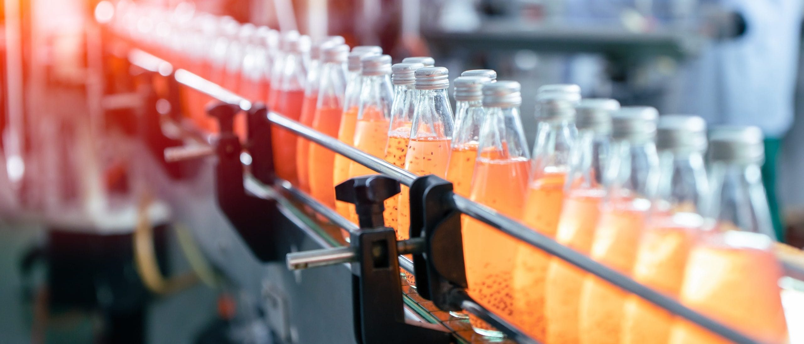 A row of glass bottles filled with orange liquid moves along a conveyor belt in a factory setting. Workers in protective clothing are visible in the background, operating machinery. The scene is brightly lit, indicating an active production line.