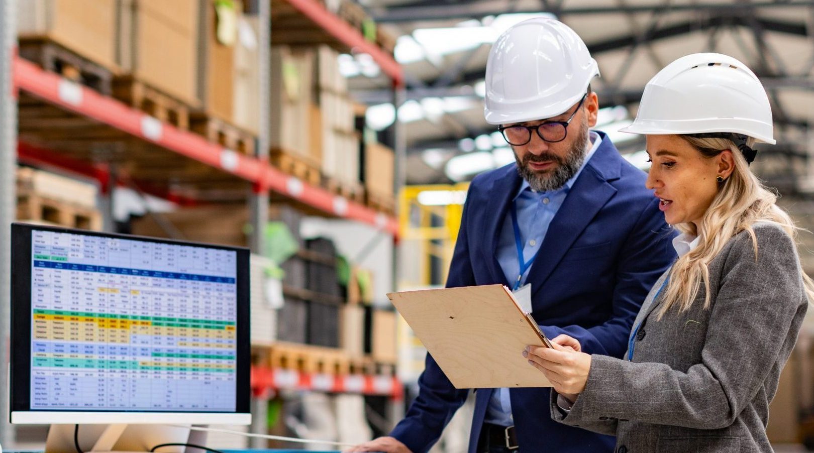 Two people in hard hats and business attire stand in a warehouse, examining a document on a clipboard. A monitor displaying data is on a nearby cart. Shelves filled with boxes and materials are in the background.